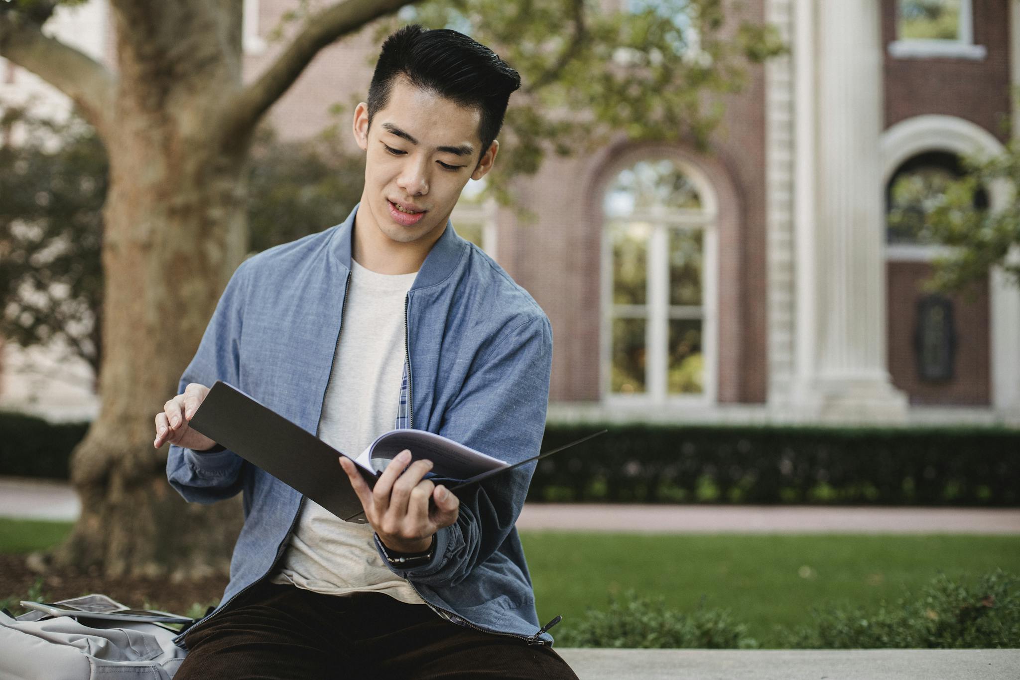 Asian man studying on a university campus bench, focusing on documents.